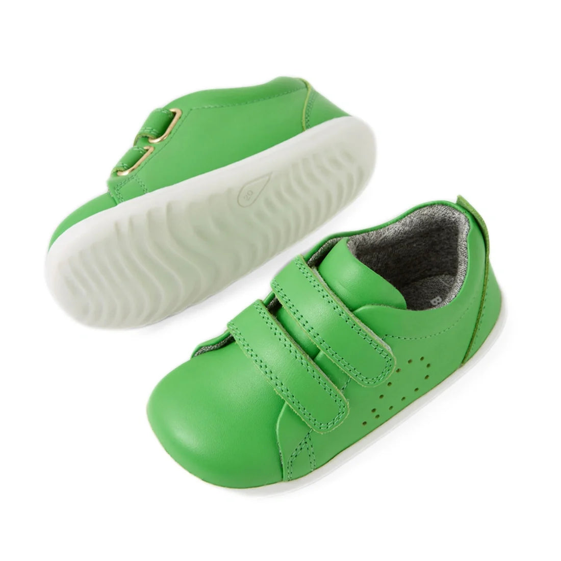 Two green Bobux Step Up Grass Court Trainer Retro Green toddler shoes with white rubber soles; one shoe displays the front featuring two Velcro straps, while the other shows off a wavy textured grip on the sole. These kids’ leather sneakers are an excelle