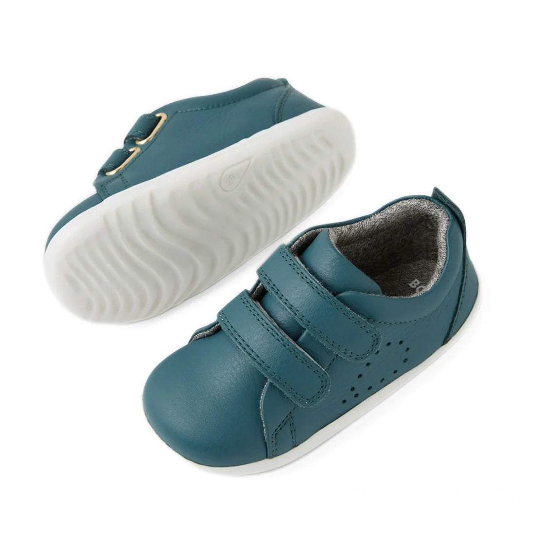 Shown on a white background, the Bobux Step Up Grass Court Trainer in Mediterranean Green is a pair of kids’ sneakers crafted from premium leather with white rubber soles, velcro straps, and decorative perforations. These stylish first walker shoes are de