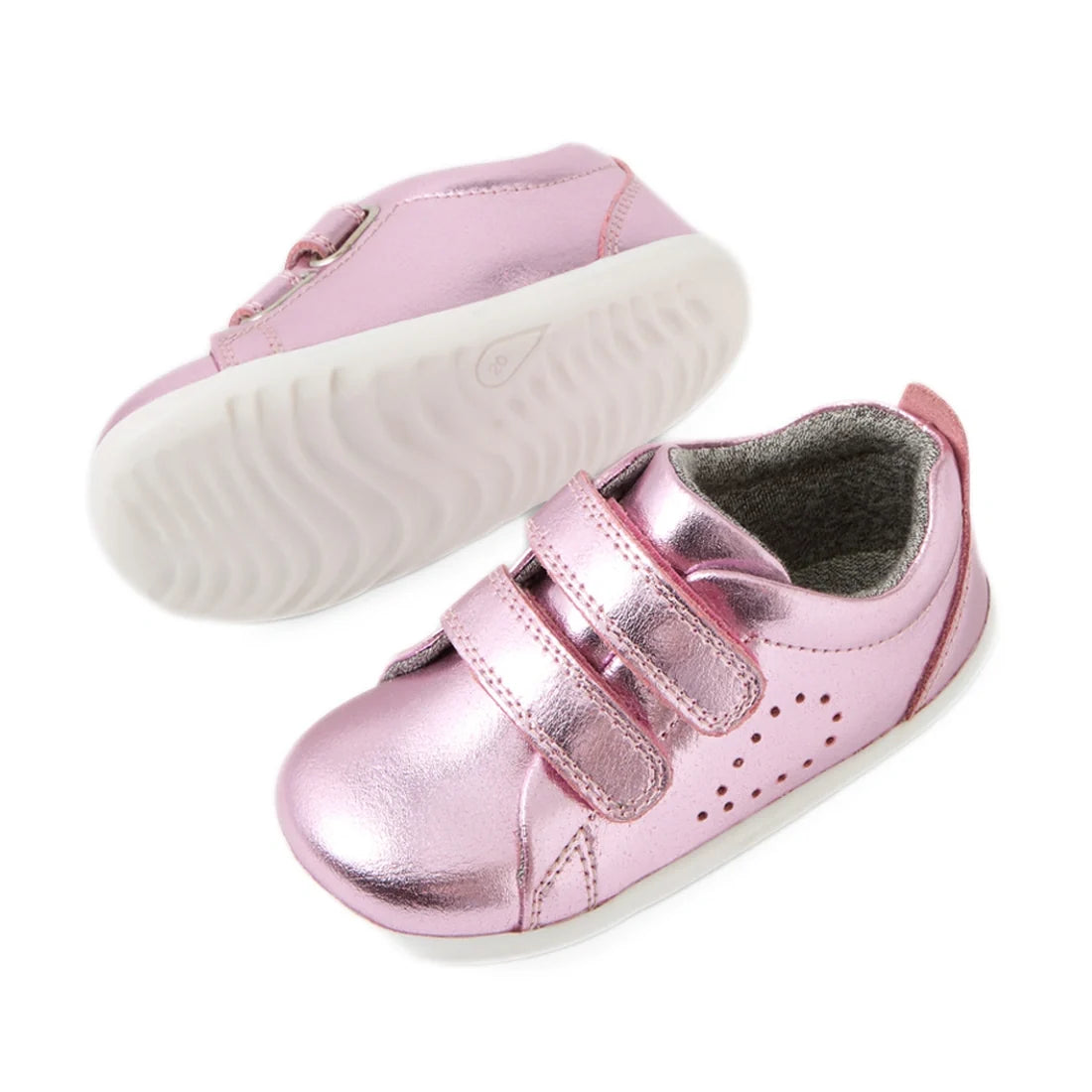 Two Bobux Step Up Grass Court Trainer Pink Metallic baby shoes with white soles and two Velcro straps are shown. Designed as ideal first walkers, one shoe is displayed upright to showcase the top and side, while the other is upside down, revealing the sol