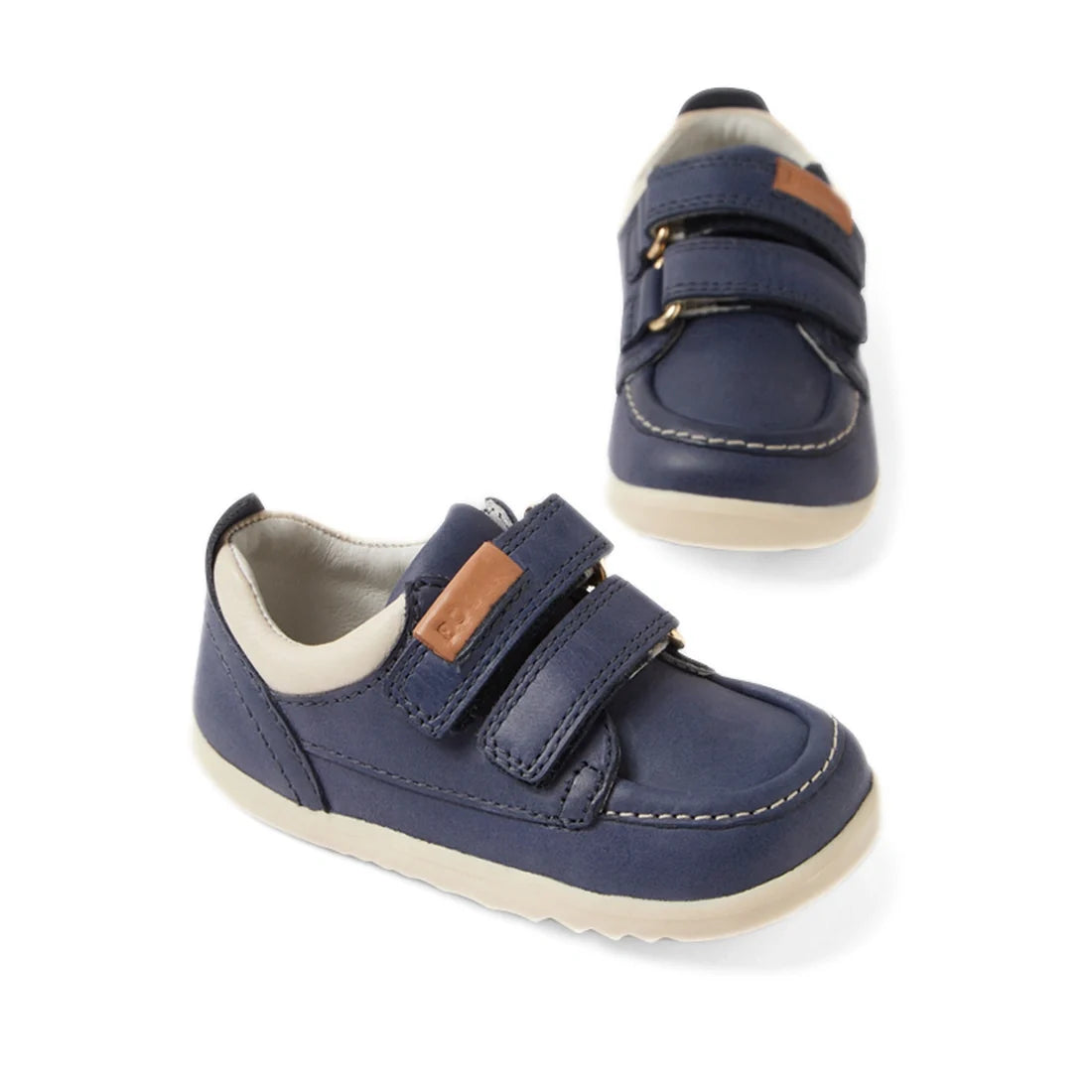 Two Bobux SU Step Up Oakley Leather Toddlers Sneakers in Vintage Indigo are displayed at different angles on a white background. These kids’ first walker shoes feature navy blue leather uppers, cream soles, a beige interior lining, two velcro straps each,