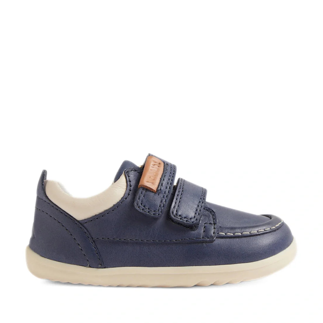 The Bobux SU Step Up Oakley Leather Toddlers Sneaker in Vintage Indigo is a stylish navy blue first walker shoe inspired by classic moccasin boat shoe design. It features two Velcro straps, a beige inner lining, and a light-colored rubber sole, finished w