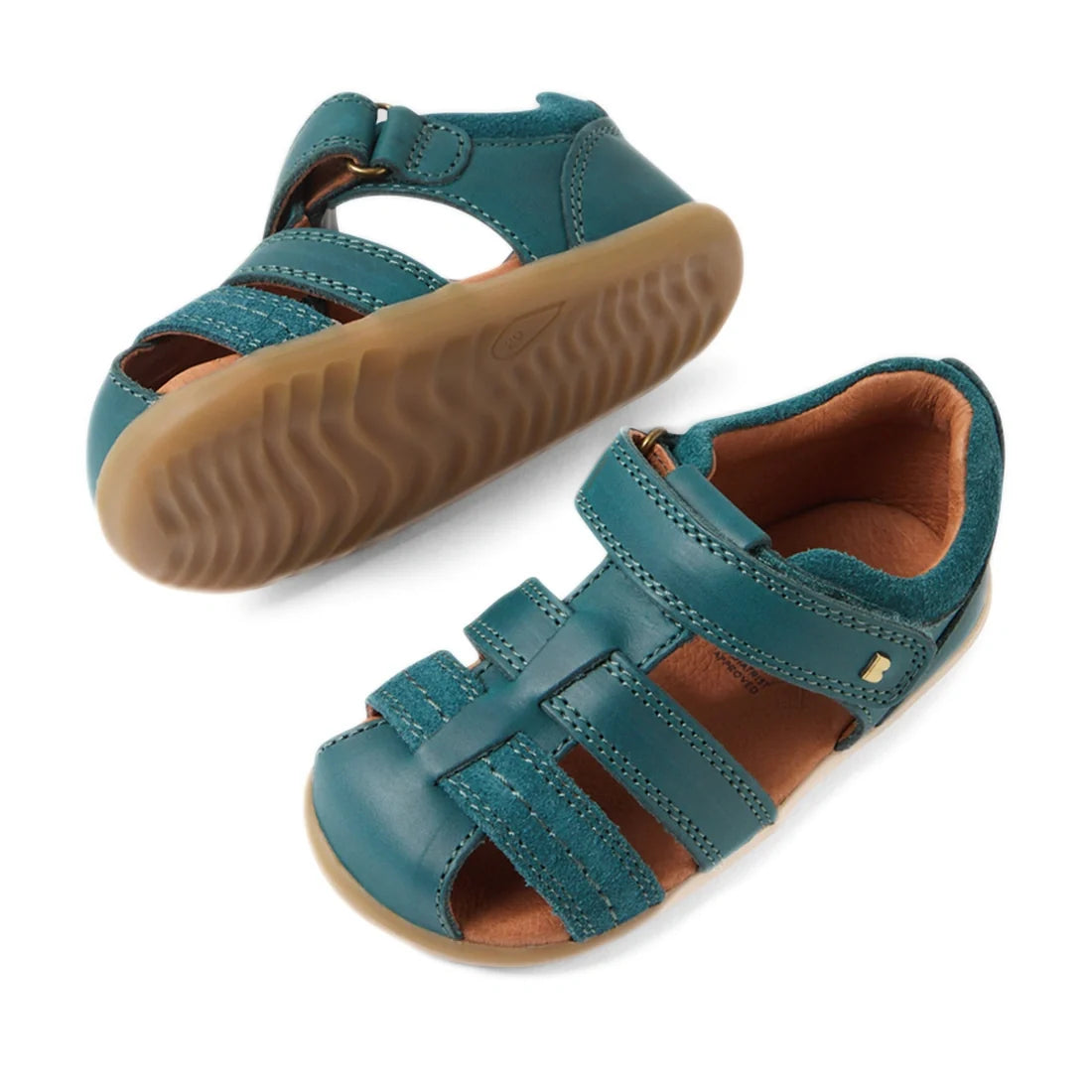 A pair of Bobux Step Up Sandal Roam Closed Toe Sandals in Mediterranean Green, featuring tan rubber soles, velcro straps, and closed toes. Crafted from durable leather, one sandal is upright while the other shows off the distinctive wavy tread pattern on 