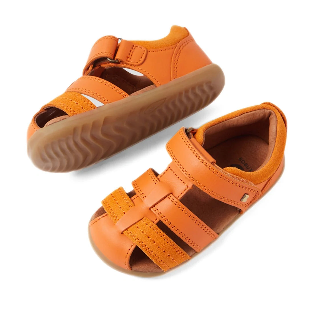 The Bobux Step Up Sandal Roam Closed Toe Sandals in Orange Peel are displayed on a white background. One sandal stands upright while the other is tilted to reveal the tread pattern, showcasing the quality and design of these toddler leather sandals with V