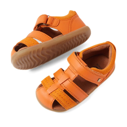 The Bobux Step Up Sandal Roam Closed Toe Sandals in Orange Peel are displayed on a white background. One sandal stands upright while the other is tilted to reveal the tread pattern, showcasing the quality and design of these toddler leather sandals with V