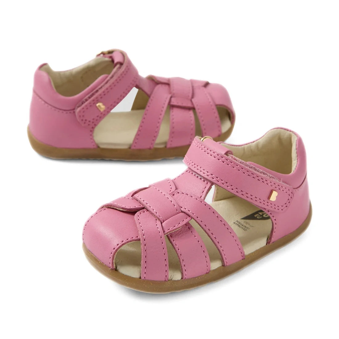 Two Bobux Step Up Sandal Skip Twist Closed Toe Sandals in cashmere rose pink are displayed on a white background—one facing forward and the other turned to the side. Featuring woven leather straps, Velcro closures, and light brown soles, these first walki