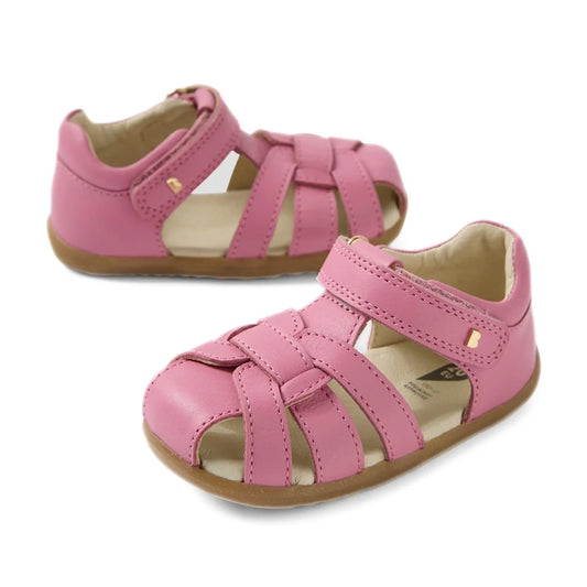 Two Bobux Step Up Sandal Skip Twist Closed Toe Sandals in cashmere rose pink are displayed on a white background—one facing forward and the other turned to the side. Featuring woven leather straps, Velcro closures, and light brown soles, these first walki