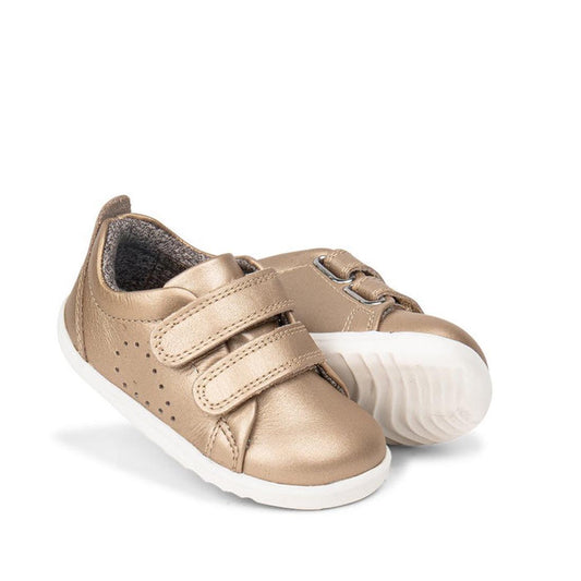 The Bobux Step Up Grass Court Trainer 728918 in gold features a leather upper, white soles, two Velcro straps, and perforated side details. One toddler sneaker stands upright while the other leans against it, displaying the sole.
