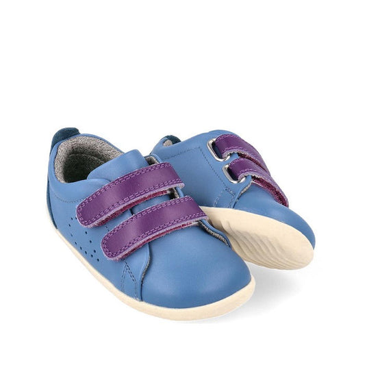 The Bobux Step Up Grass Court Trainer Elemental Blue 734205 features a blue leather upper, off-white soles, and two purple Velcro straps per shoe, displayed against a white background.