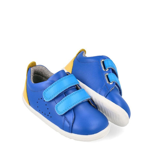 The Bobux Step Up Grass Court Trainer Azzure Blue 734207 features blue uppers, light blue Velcro straps, white soles, and yellow heel accents, shown on a white background.