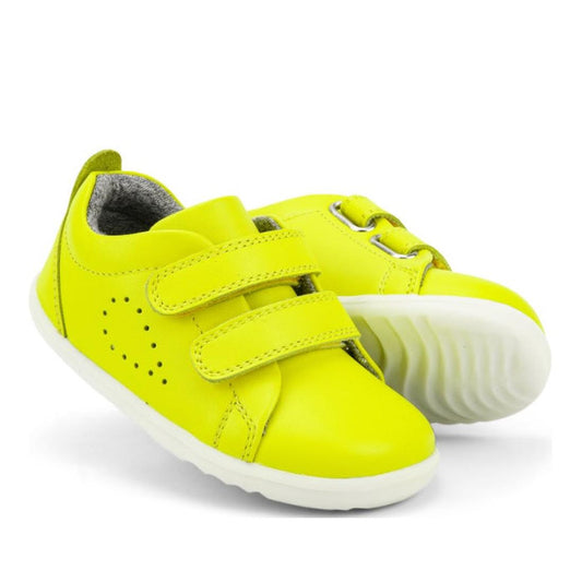 The Bobux Step Up - Grass Court Toddlers Sneaker 728941 in neon yellow features bright leather, white soles, and two adjustable straps. Displayed with one shoe slightly overlapping the other.