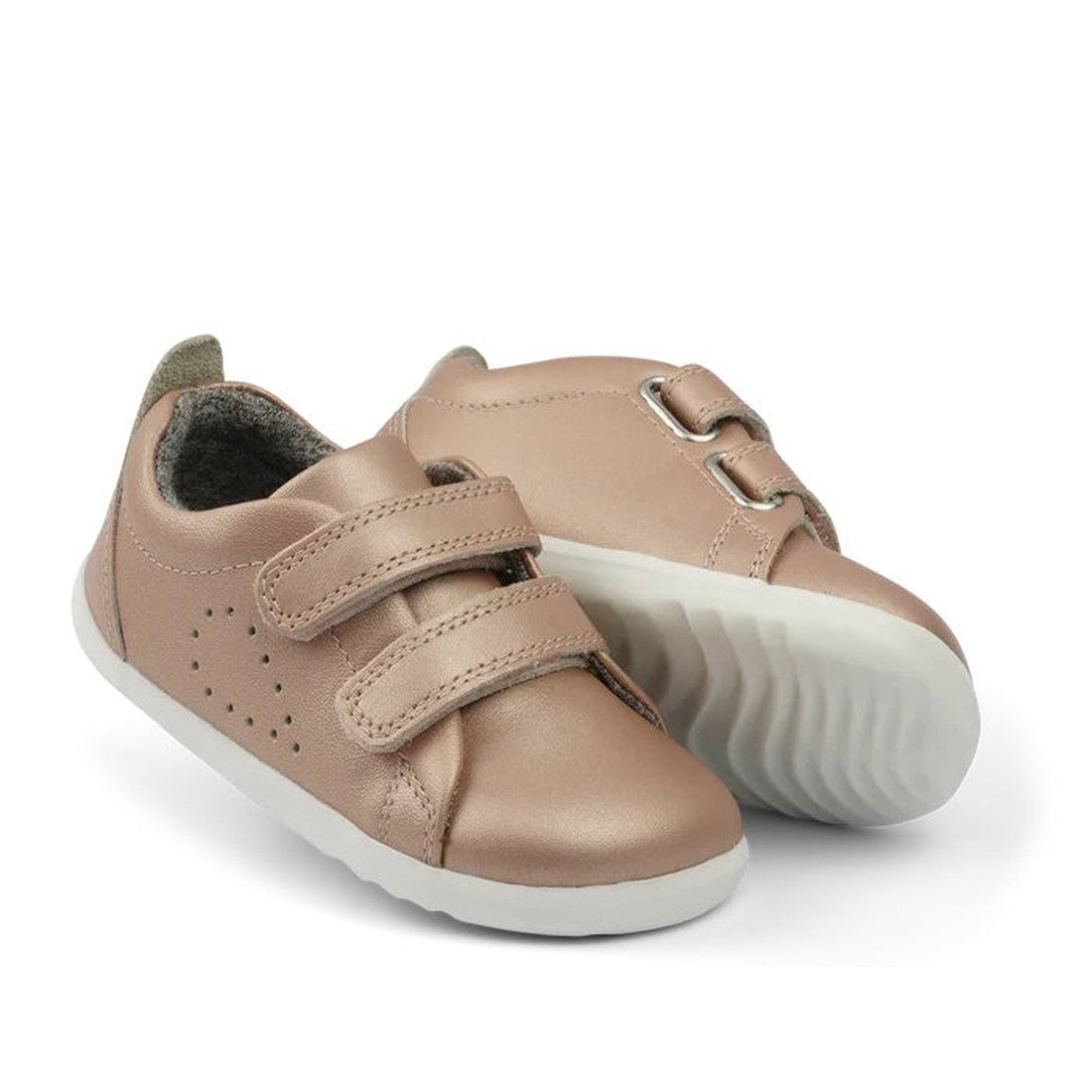 The Bobux Step Up Grass Court Trainer 728926 in Rose Gold is a pair of metallic leather toddler shoes with white soles, two hook-and-loop straps, and side perforations—ideal for first walkers. One shoe stands upright, the other lies on its side.