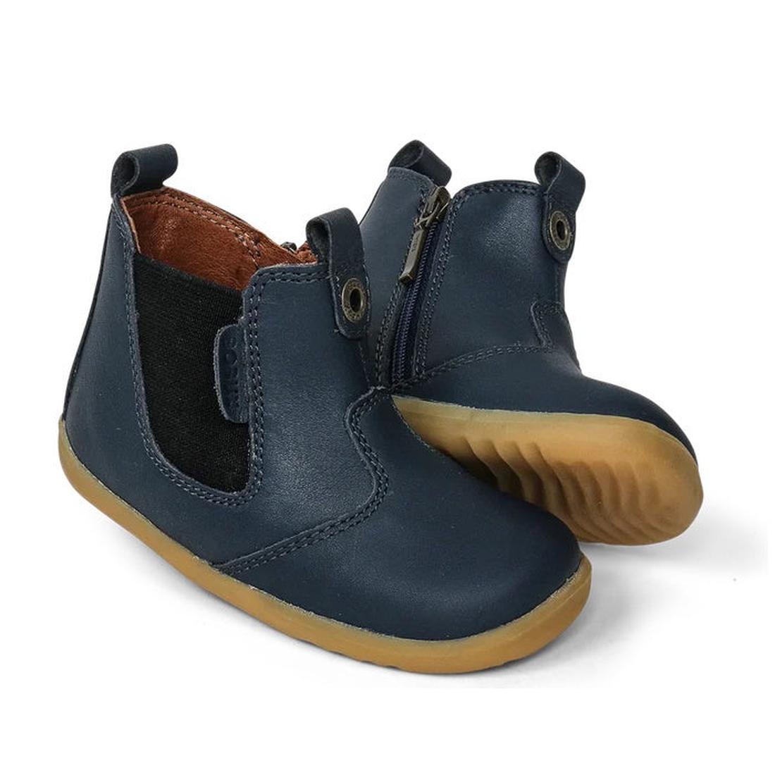 The Bobux Step Up Jodphur Boot Navy 721929A features navy blue leather, tan rubber soles, black elastic side panels, and a side zipper. These unisex toddler ankle boots are ideal as first walkers; one boot stands upright, the other shows the sole.