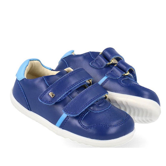The Bobux Step Up SU Riley Sneaker Blueberry is a unisex children's blue sneaker with white soles, light blue accents, and two hook-and-loop velcro straps for easy fastening.