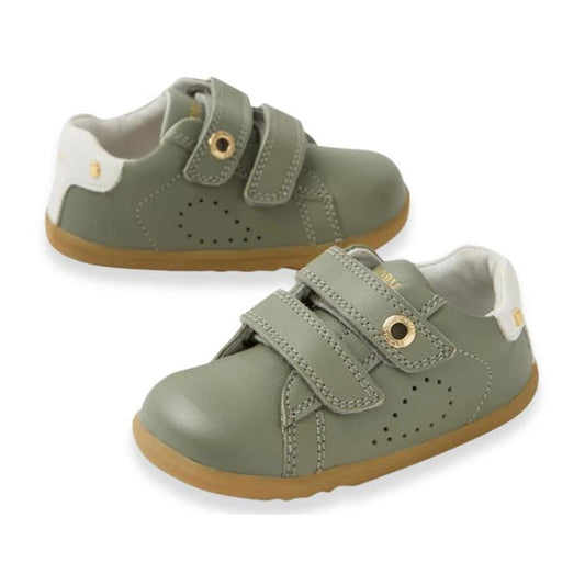 The Bobux SU Step Up Maverick Leather Toddlers Sneaker in Vetiver Green features olive green leather, tan rubber soles, white heel accents, gold eyelets, and two adjustable Velcro straps per shoe.