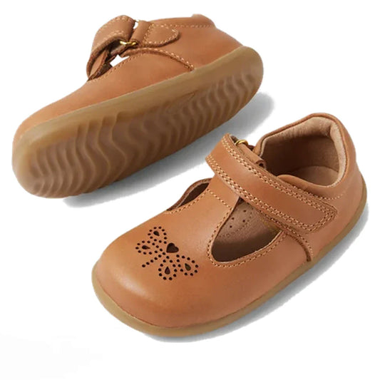 The Bobux Step Up T-Bar Rosie First Walker Shoe in Caramel Tan is a light brown toddler leather flat with Velcro straps, rubber soles, and a butterfly cut-out with heart on the toe—ideal for first walkers. One shoe stands upright, one lies on its side.