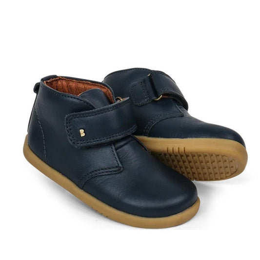 The Bobux Iwalk IW Desert Boot Navy 625206A is a pair of children’s navy blue leather desert boots with brown rubber soles and hook-and-loop strap closures, designed for growing feet and shown against a white background.