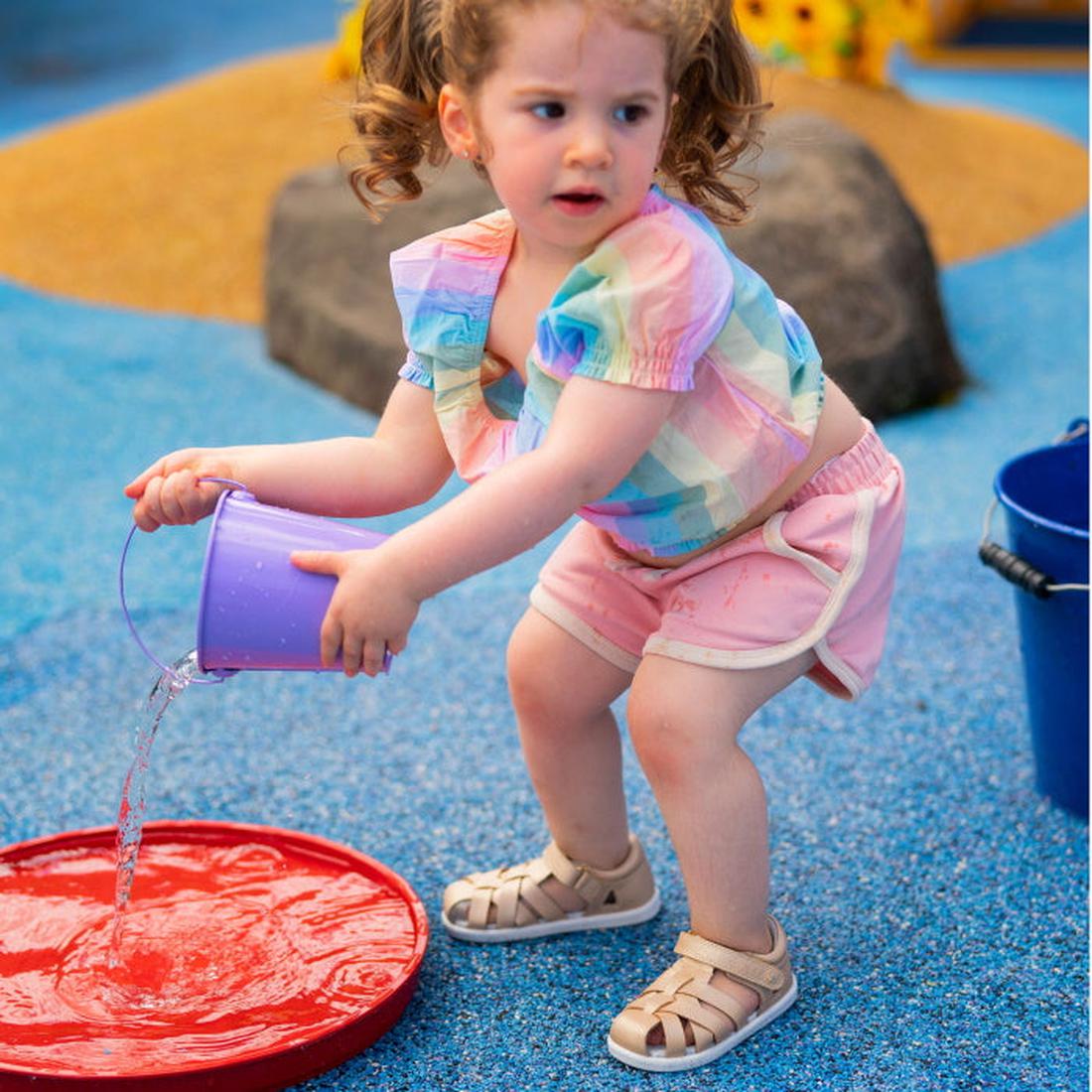 A young girl with curly hair, wearing a colorful shirt, pink shorts, and Bobux Step Up SU Tropicana II Pale Gold 732311 sandals, crouches at a splash pad as she pours water from a purple bucket onto the red surface., 9420068529915, 9420068529922, 94200685