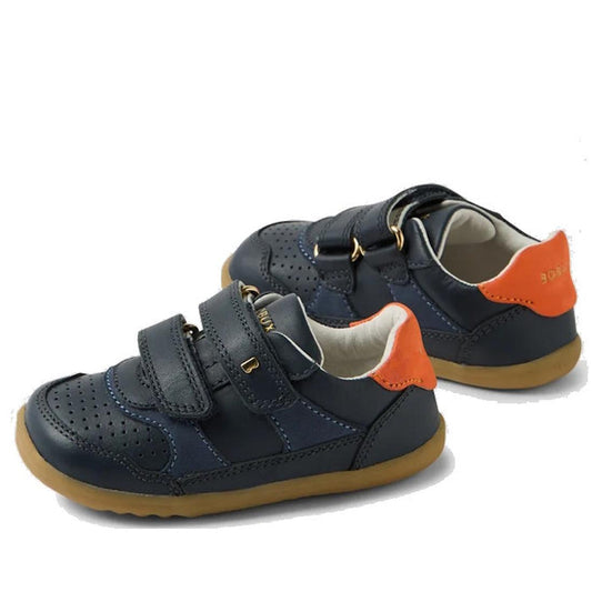 The Bobux SU Step Up Hunter Leather Toddlers Sneaker in navy features orange heel accents, tan rubber soles, double Velcro straps, perforated toe detailing, and the BOAR brand on the straps and heel—perfect for first walkers.