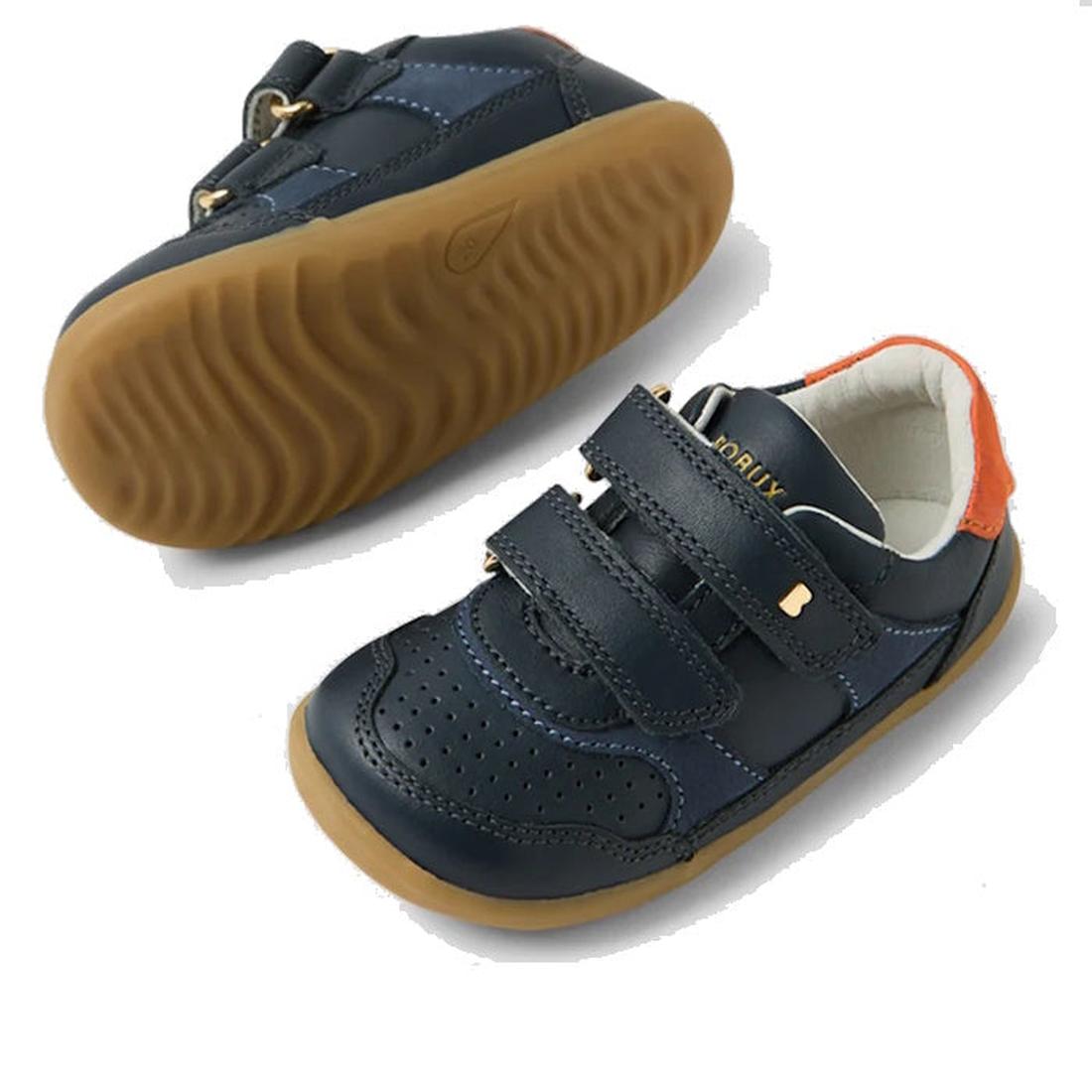 The Bobux SU Step Up Hunter Leather Toddlers Sneaker in navy features brown rubber soles, double Velcro straps, perforated toes, and orange heel accents. One shoe is upright; the other is shown from the side., 9420074833853, 9420074833860, 9420074833877