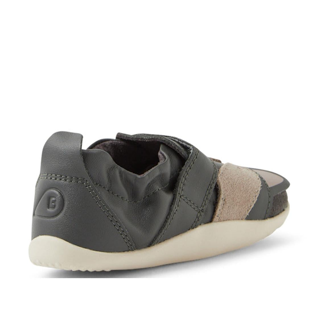 The Bobux XP Xplorer Scrunch First Walking Shoes in Smoke Grey are pre-walker toddler shoes in dark gray with beige suede accents, shown from a rear-side angle. They feature an adjustable strap, flexible material, and a light-colored textured sole., 94200