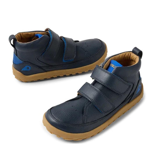 The Bobux Kid+ Trek Leather Boot Navy Blue 853801 features a barefoot-shaped toe box, tan rubber soles, blue accents, and two Velcro straps. Shown in navy blue on a white background.