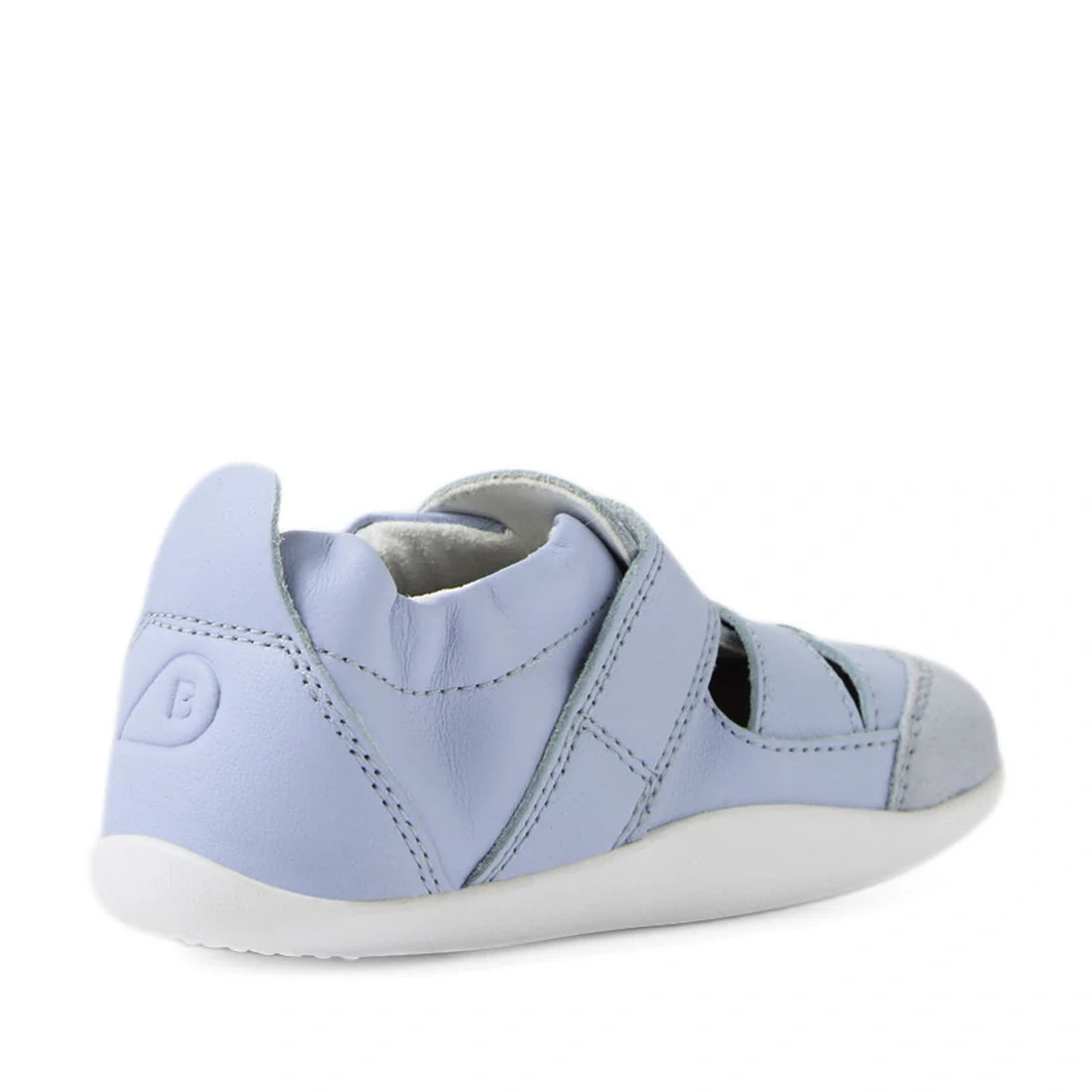 The Bobux XP Explorer Maris First Walker Sandal in Skyway Blue is a light blue children’s shoe featuring a soft, flexible sole, side cutouts, and a closed toe inspired by classic fisherman sandals—shown here from the back left side. The upper combines lea