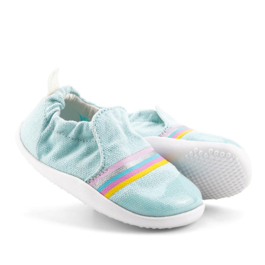 The Bobux Xplorer Scamp Crawler Aqua Silver Rainbow 501709 Canvas are vegan-friendly, light blue slip-on summer shoes for toddlers, featuring elastic openings and pastel rainbow stripes with white soles—perfect for little ones on the go.
