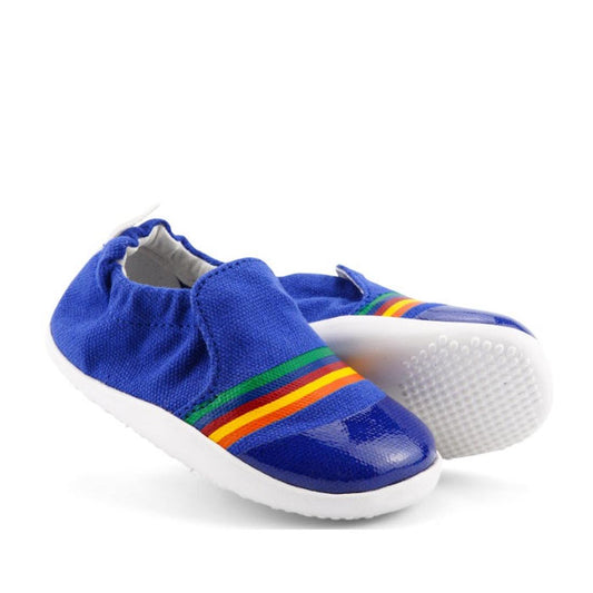 The Bobux Xplorer Scamp Crawler Blueberry Red Rainbow 501711 Canvas shoes feature blue canvas, white flexible soles, elastic heels, red/yellow/green side stripes, and glossy blue accents on the toe. Perfect for baby's first steps.