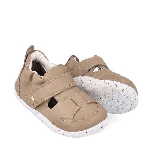 The Bobux XP Explorer Beau First Walker Sandal Atmosphere 502301 is a pair of beige toddler shoes with flexible, white speckled rubber soles, velcro straps, and top cutout details, shown on a white background.