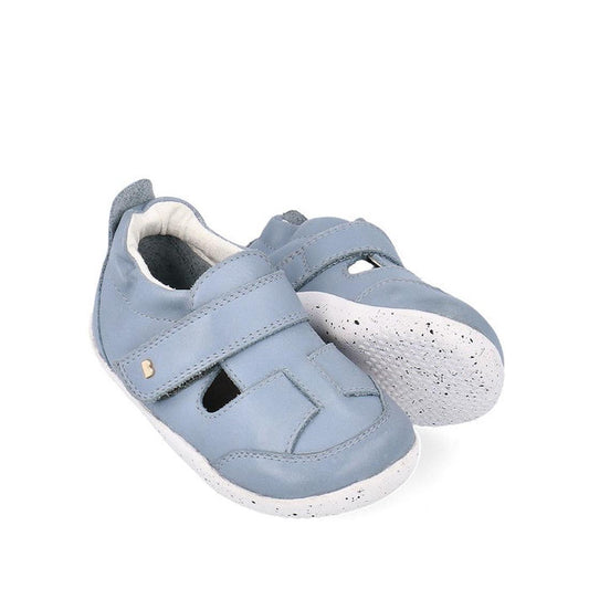 The Bobux XP Explorer Beau First Walker Sandal 502303 features light blue uppers, white flexible soles, Velcro straps, and front cutout details, displayed against a white background.