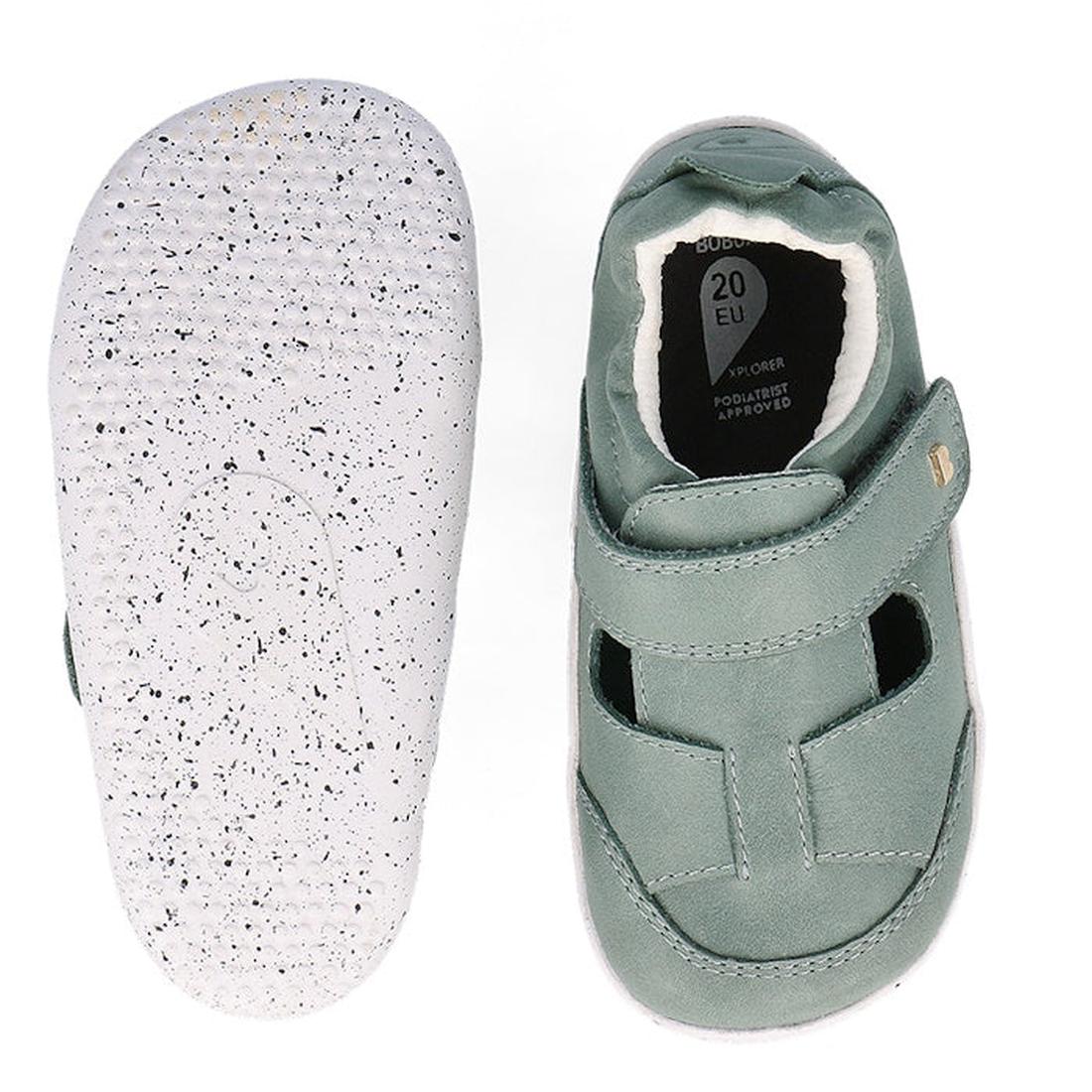 The Bobux XP Explorer Beau First Walker Sandal in Sage Green features white, speckled rubber soles, a hook-and-loop strap, and a flexible sole with a dotted tread pattern. One shoe is shown top-down; the other displays the sole., 9420075784499, 9420075783