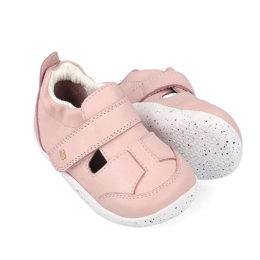 The Bobux XP Explorer Beau First Walker Sandal Blossom Pink 502305 features light pink uppers, velcro straps, white flexible soles, a gold B logo, and cute cut-out details on top. Displayed against a white background.