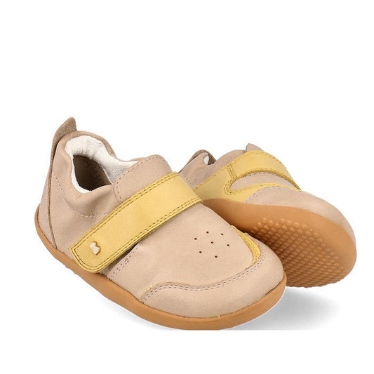 The Bobux XP Explorer Ripple First Walker Sneaker Beige 502502 features beige uppers, light brown flexible soles, yellow hook-and-loop straps, toe ventilation holes, and is an ideal first walker shoe for toddlers.