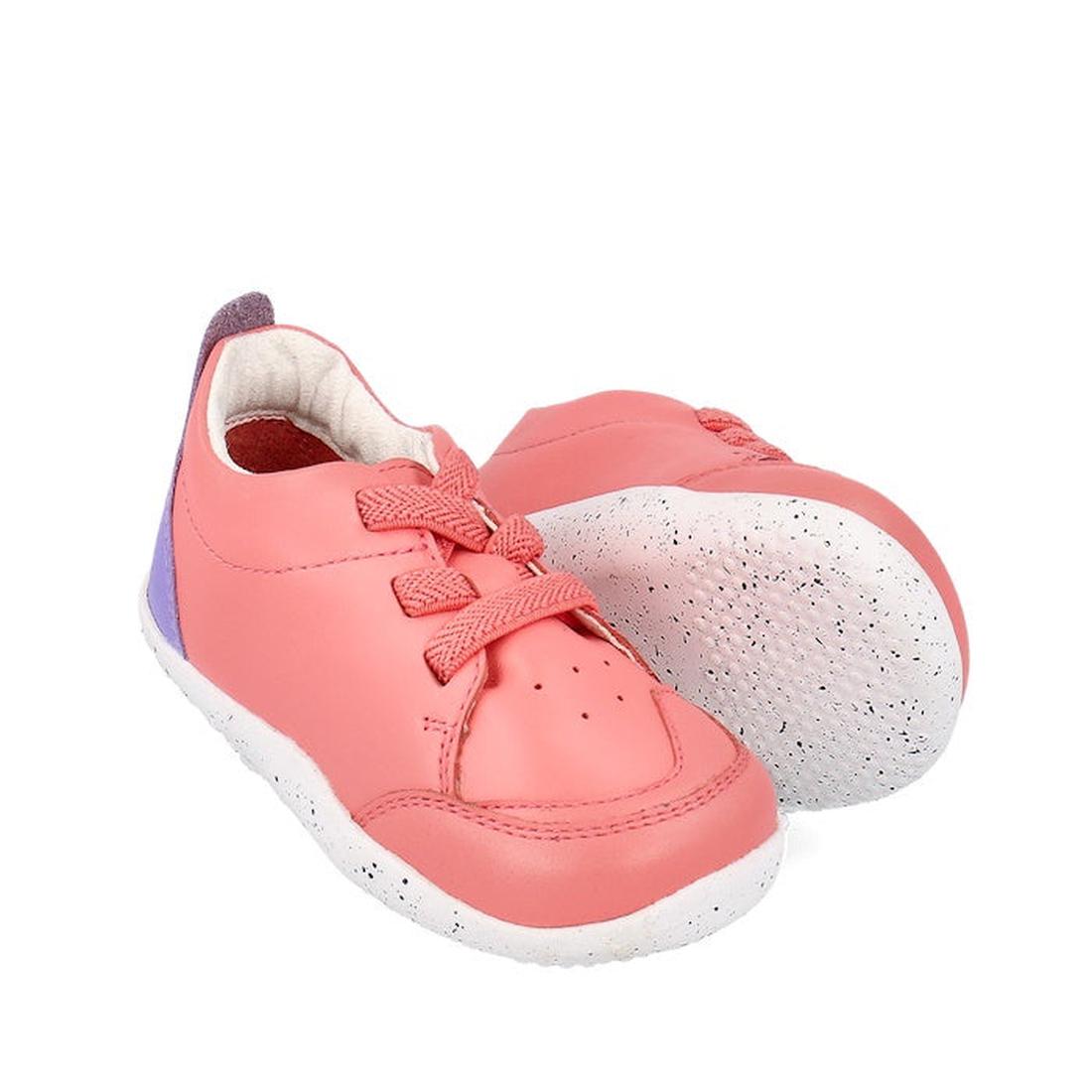 The Bobux XP Explorer Sport First Walker Sneaker Ocean Coral 502204 is a pair of pink toddler shoes with purple heel accents, white speckled soles, and pink laces. One shoe stands upright while the other shows its flexible sole for comfort.