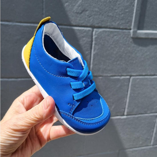 A hand holds the Bobux XP Explorer Sport First Walker Sneaker Azzure Blue 502206, featuring bright blue laces, a white sole, and yellow heel accent—perfect for first walkers—in sunlight against a gray brick wall.