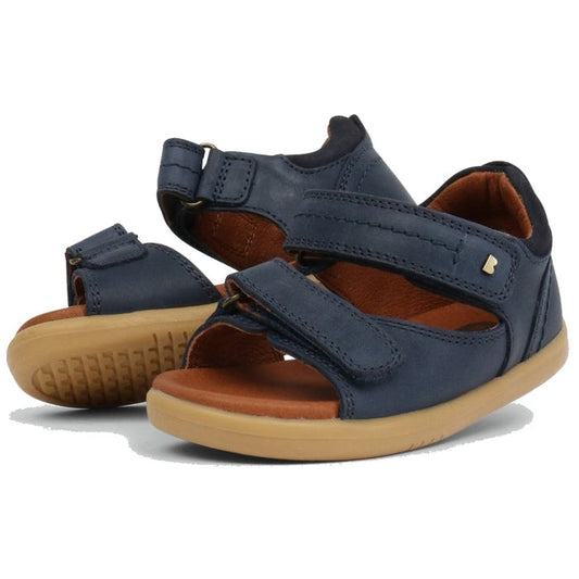 The Bobux IW Iwalk Driftwood Sandal Navy Open Toe 633601A is a children's navy sandal with two adjustable Velcro straps, tan rubber soles, closed heels, open toes, and a small metallic side logo.