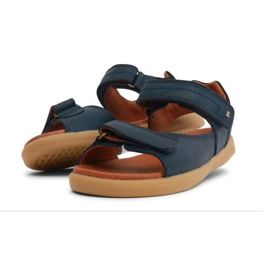 A pair of Bobux Kid+ Plus Driftwood Navy Open Toe Sandals (style 833501AA) for kids, featuring navy and tan leather with adjustable Velcro straps, displayed at an angle on a white background.