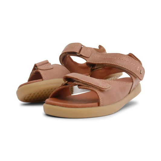 The Bobux Kid+ Plus Driftwood Caramel Open Toe Sandal 833504A features a tan leather upper, adjustable double strap, and light brown rubber sole. Displayed on a white background, one sandal slightly overlaps the other.