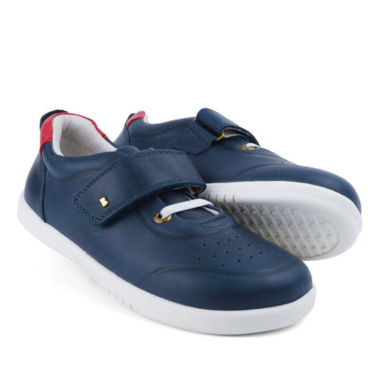 The Bobux Kids + Plus Ryder Navy Velcro Trainers (835602) feature blue leather, white soles, gold eyelets, a velcro strap design, and a small red heel accent, shown on a white background.