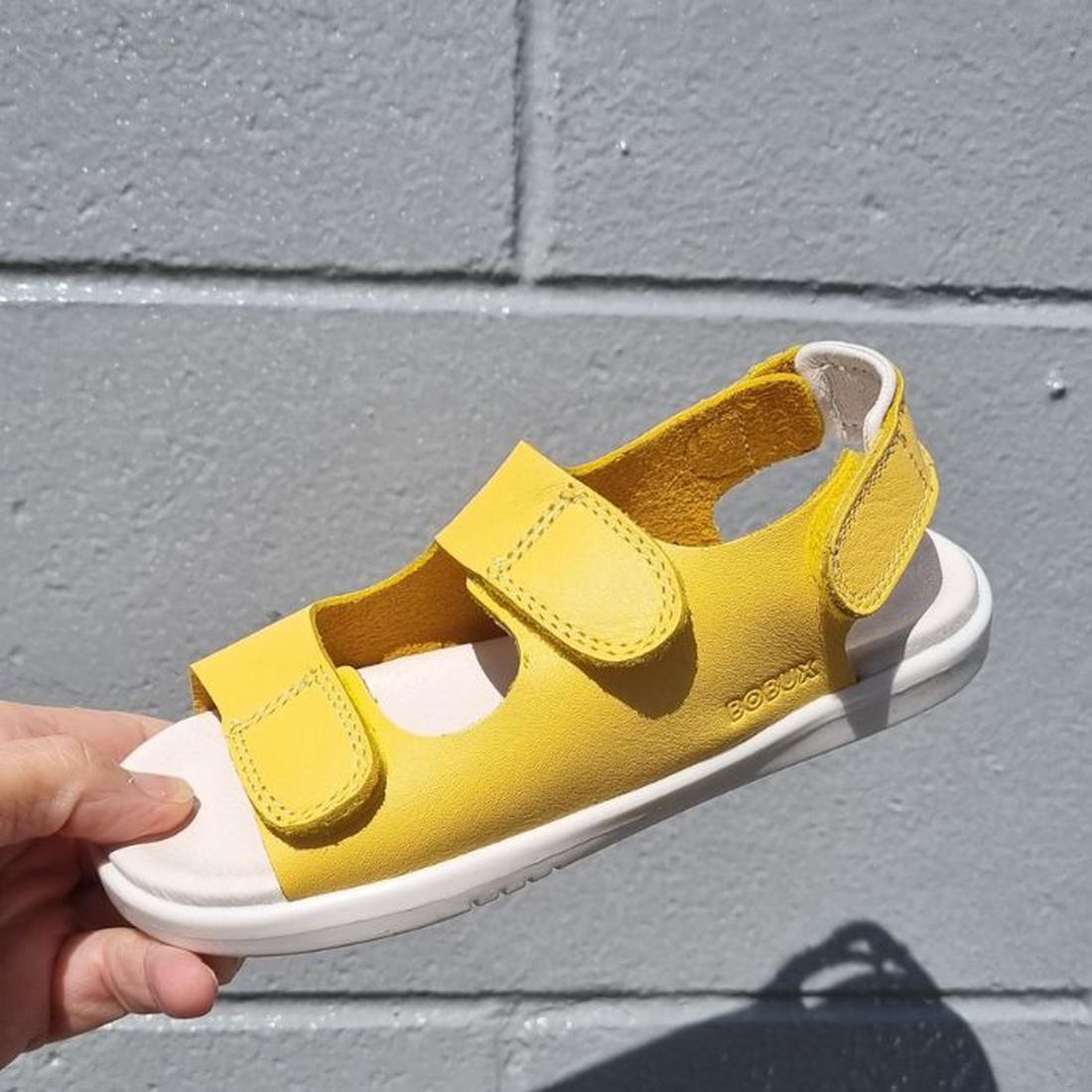 A hand holds the Bobux Kids Plus KP Topia Quick Dry Sandal in Pineapple Yellow, featuring two Velcro straps and a white sole, set against a gray concrete block wall background.