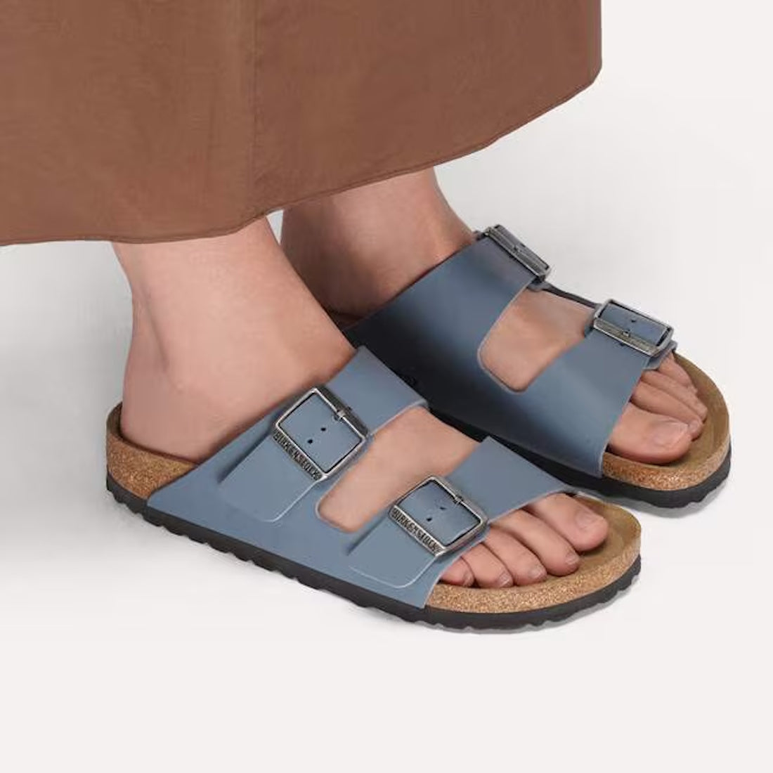 A person stands on a light surface wearing a brown skirt and BIRKENSTOCK Arizona Birko Flor Balsat Grey Regular Width 2 Strap Sandals with supportive cork-latex footbeds., 51737, 51738, 51739, 4067707675807, 4067707675845, 4067707675883, 4067707675920, 40