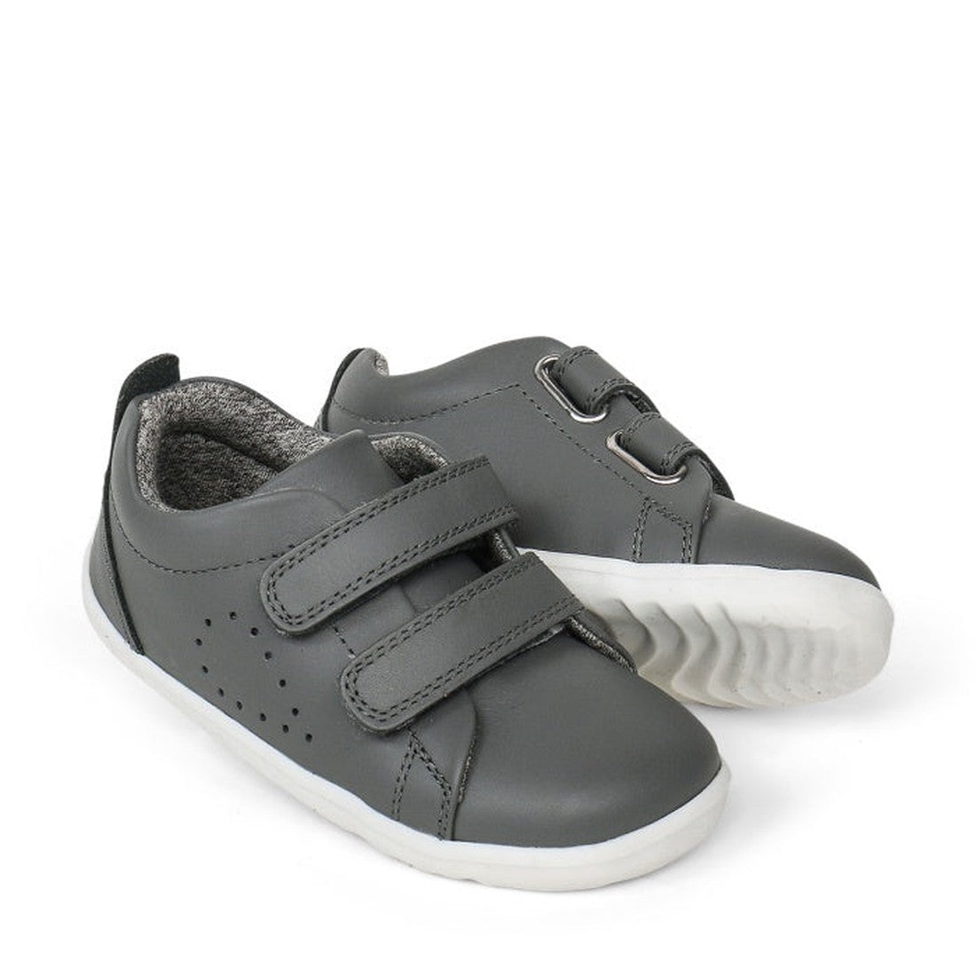 A pair of Bobux Step Up First Shoes Grass Court Trainer 728913 in Smoke Grey, featuring soft leather, white soles, and Velcro straps. One shoe is slightly behind and angled to display the side and front—ideal for first walkers.