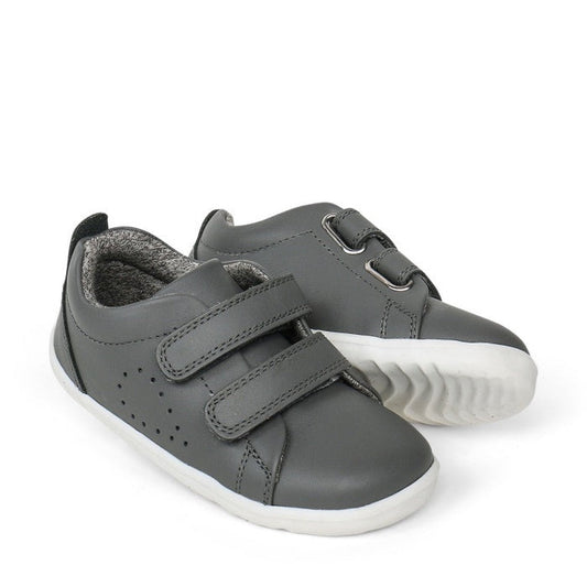 A pair of Bobux Step Up First Shoes Grass Court Trainer 728913 in Smoke Grey, featuring soft leather, white soles, and Velcro straps. One shoe is slightly behind and angled to display the side and front—ideal for first walkers.