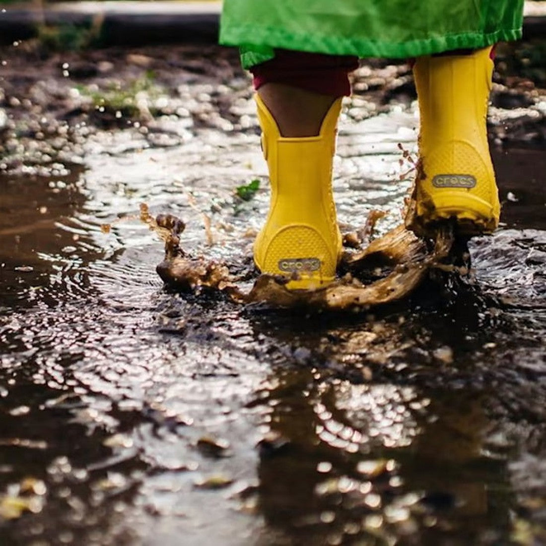 A child in green pants wearing Crocs Kids Handle It Rain Boot Yellow jumps into a muddy puddle, sending water and mud flying. The photo highlights the dynamic motion and playful energy of the moment., 883503861213, 883503861220, 883503861237, 883503861244