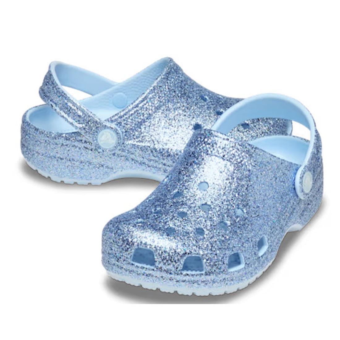 The Crocs Kids Classic Chunky Glitter Clogs are light blue, feature glitter, ventilation holes, and adjustable heel straps, inspired by classic Crocs. Displayed on a white background.