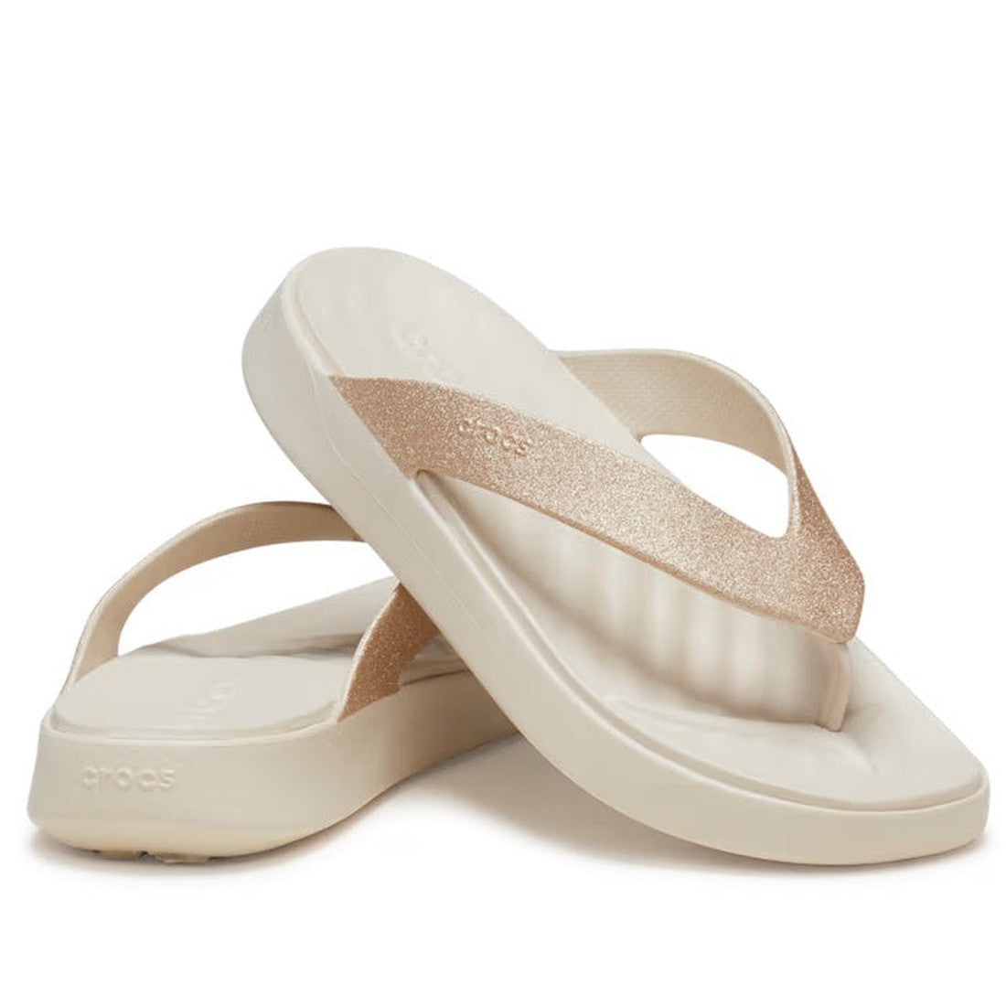 A pair of Crocs - Womens Getaway Glitter Strappy Flip Flop Sandals in Stucco, featuring metallic rose gold straps and a cushioned footbed with Free Feel Technology textured sole. One sandal stands upright while the other is on its side.