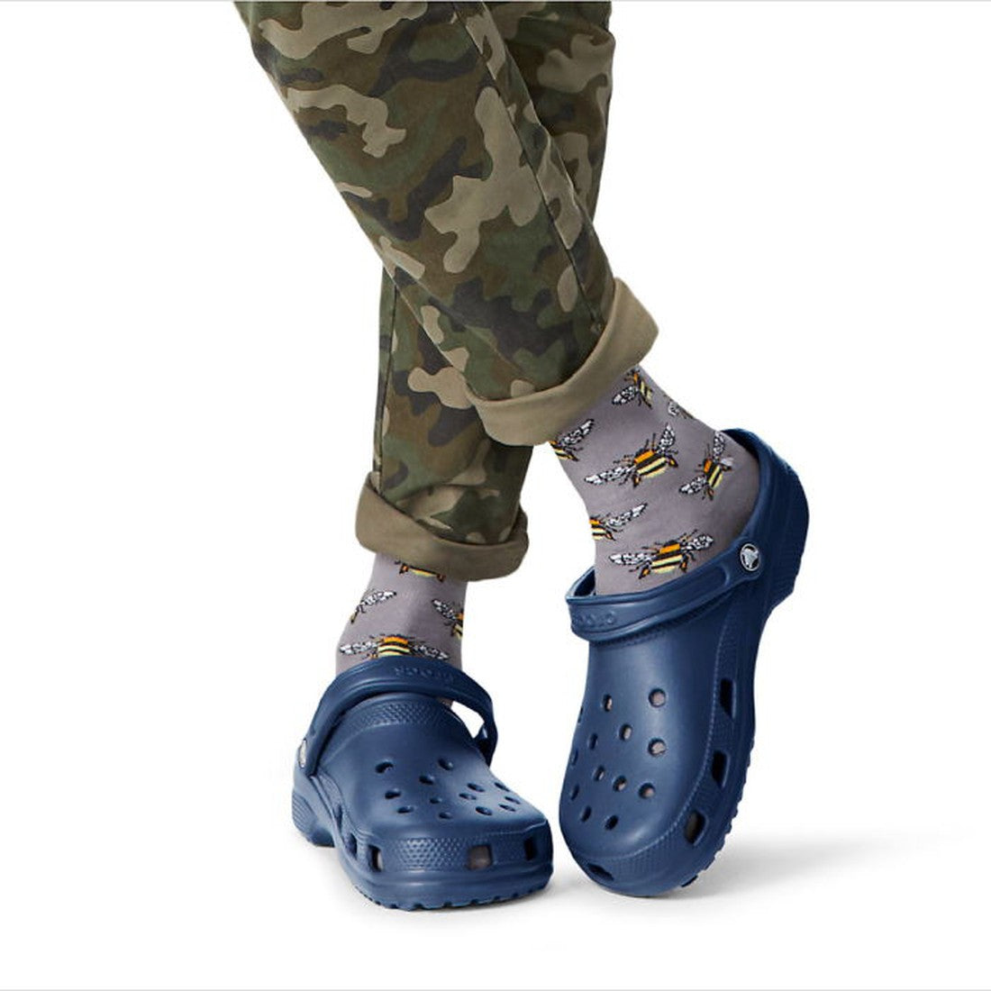 Person wearing camouflage pants and gray socks with yellow bees, paired with Crocs - Classic Clog Navy Adults womens & mens. Legs are crossed, highlighting the unisex style and comfort—only the lower half is shown., 841158002696, 841158002702, 84115800271