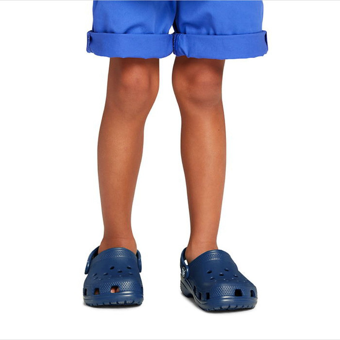 A child wearing blue shorts and Crocs Kids - Classic Clog K Navy Youths, crafted from soft Croslite for all-day comfort, stands facing forward with legs slightly apart against a white background., 191448735002, 191448735019, 191448735026, 191448735033, 19