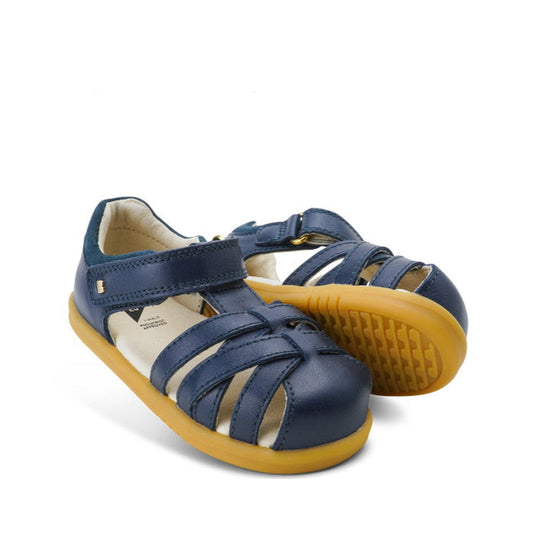 A pair of Bobux Step Ups Cross Jump Navy Closed Toe Sandal unisex with adjustable velcro straps and yellow rubber soles is displayed on a white background.