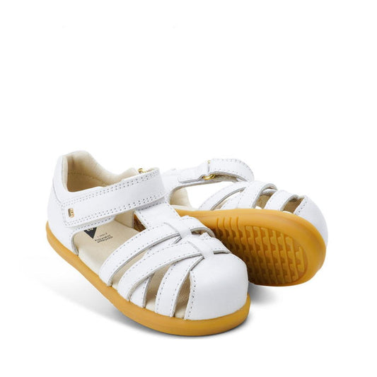 The Bobux Step Ups Cross Jump White 731106 Closed Toe Sandal features a white leather upper, closed toes, open sides, tan rubber soles, and Velcro straps. One sandal stands upright while the other leans to show the sole.